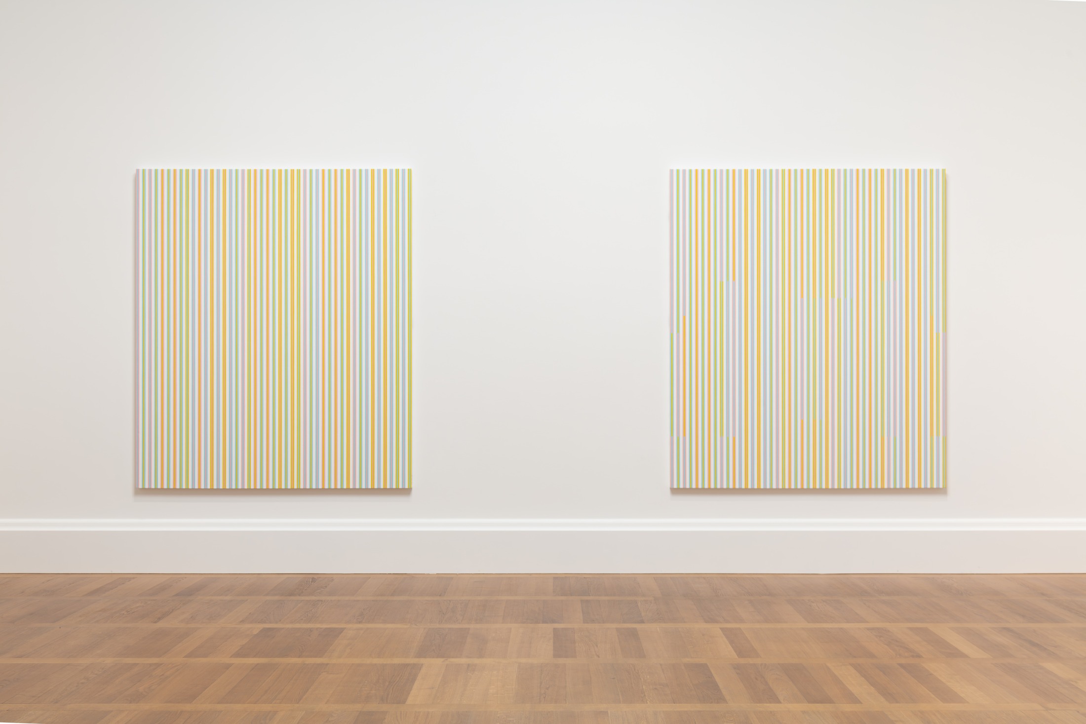 A gallery photo of two Bridget Riley works with vertical colored bands on a white wall.